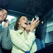 Icon 1461814354 9 20 12 businesswoman in a taxi applying lipstick sat next to a businessman using a mobilephone